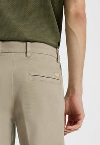 Beige broek met een gestructureerde afwerking, voorzien van een discreet achterzakje en een klein logo-opdruk. Gepaard met een gestructureerde donkergroene top.