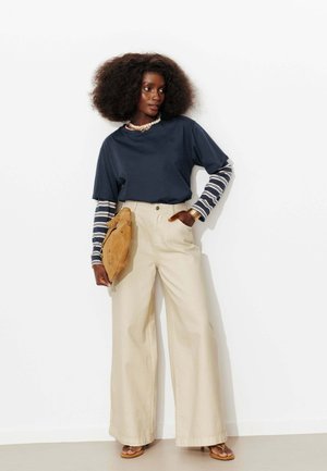 Femme aux cheveux bouclés portant des manches longues superposées bleu marine et rayées, un pantalon beige à jambes larges, des sandales, et tenant une pochette beige contre un mur blanc.