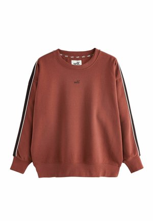 Junge Frau mit geschlossenen Augen lächelnd, trägt braunen Pullover und Hose mit weißen Streifen und "self."-Text auf dem Pullover.