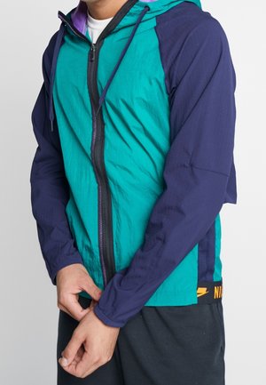 Persona con chaqueta de cremallera en tonos turquesa y azul marino con capucha, ajustando la manga izquierda, de pie frente a un fondo claro y liso.