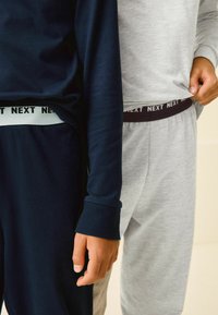 Kaksi paria oleskeluasuja: tummansininen pitkähihaisesta paidasta ja housuista, harmaasta paidasta ja housuista. Ominaisuuksia ovat logovöiden, ribattujen hihansuiden ja pehmeiden tekstuurien käyttö.
