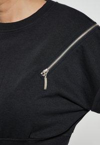Felpa nera in cotone, con una tasca a zip diagonale sulla spalla sinistra dotata di dettagli metallici e colletto a costine.