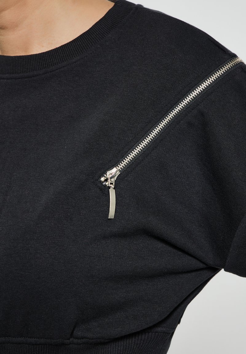 Felpa nera in cotone, con una tasca a zip diagonale sulla spalla sinistra dotata di dettagli metallici e colletto a costine.