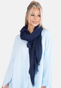 Écharpe texturée bleu marine drapée lâchement autour du cou, avec des bords effilochés, associée à une chemise bleu clair à boutons.