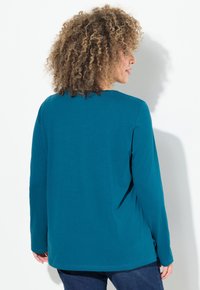 Türkises Langarmshirt aus weichem Stoff, mit rundem Halsausschnitt und lockerer Passform, sichtbaren Nähten und seitlichen Schlitzen am Saum.