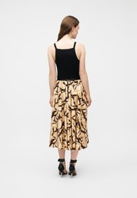 Γυναίκα με την πλάτη, φοράει μαύρο αμάνικο τοπ, μπεζ midi φούστα με μαύρο αφηρημένο μοτίβο και μαύρα σανδάλια με ψηλό τακούνι.