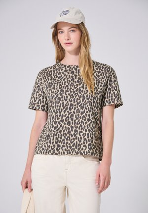 Femme portant un t-shirt imprimé léopard, un pantalon beige clair, une casquette blanche avec texte et des boucles d’oreilles créoles dorées, debout devant un fond uni.
