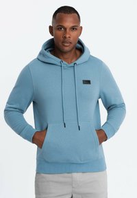 Sudadera azul claro con un bolsillo canguro, cordón ajustable, puños acanalados y un pequeño parche negro con logo en el pecho. Tejido suave.