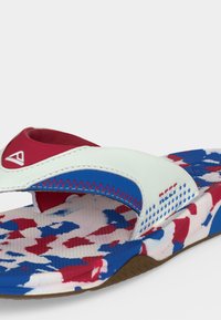 Κοντινό πλάνο ενός πολύχρωμου σανδάλιου slide με πάτο καμουφλάζ σε κόκκινο, άσπρο και μπλε και λευκό λουράκι με μπλε και κόκκινες ραφές.