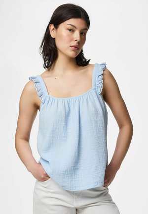 Jeune femme aux cheveux foncés portant un haut sans manches bleu clair avec des bretelles volantées et un pantalon blanc, debout les mains dans les poches.