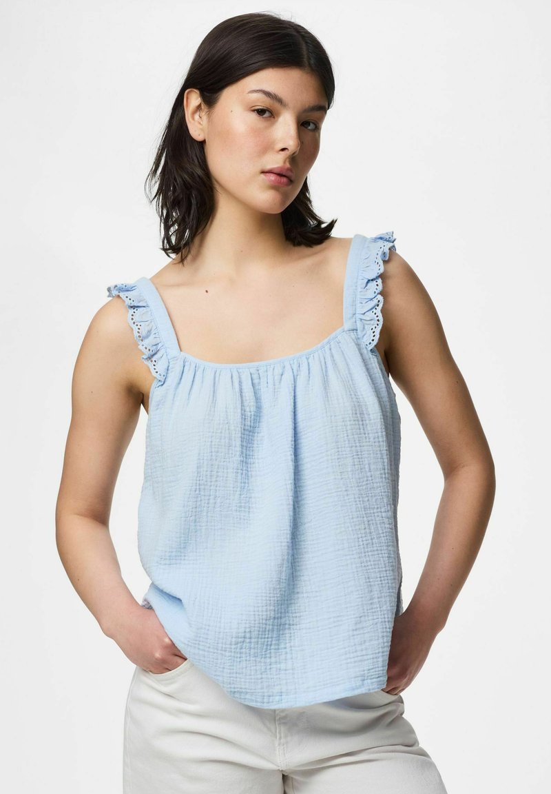 Jeune femme aux cheveux foncés portant un haut sans manches bleu clair avec des bretelles volantées et un pantalon blanc, debout les mains dans les poches.