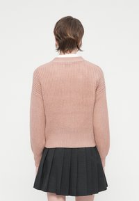 Osoba z krótkimi brązowymi włosami, odwrócona plecami, w różowym swetrze dzianinowym i ciemnej plisowanej spódnicy, na jednolitym tle.