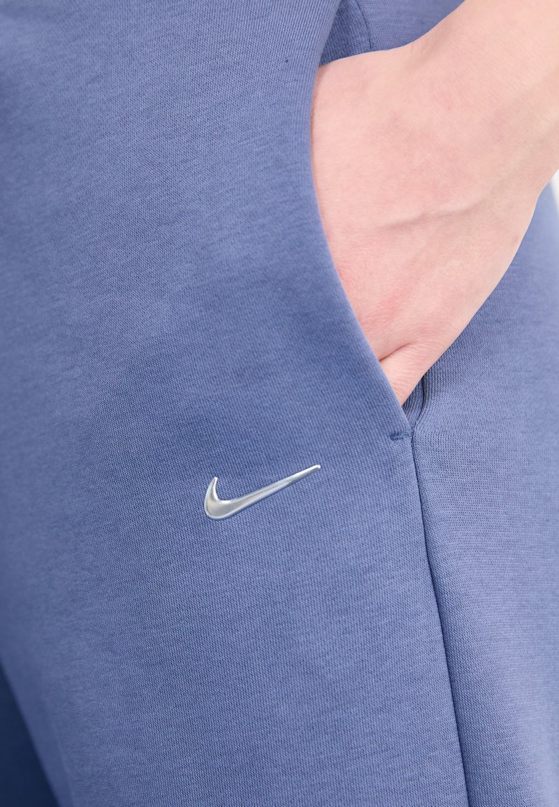 Nike Jogginghosen aus weichem blauen Stoff mit glatter Textur. Zu den Merkmalen gehören ein silbernes Logo in der Nähe der Tasche und eine geräumige Seitentasche.