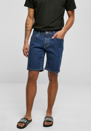Sötét kék denim rövidnadrág, laza szabással, öv hurkokkal, öt zsebbel és felhajtott szegéllyel. Fekete pántos papuccsal viselve.