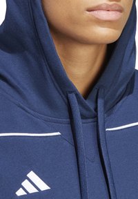 Marineblaue Kapuzenjacke mit Kordelzug, ausgestattet mit weißen Akzentstreifen und einem Adidas-Logo. Hergestellt aus weichem, strukturiertem Stoff.
