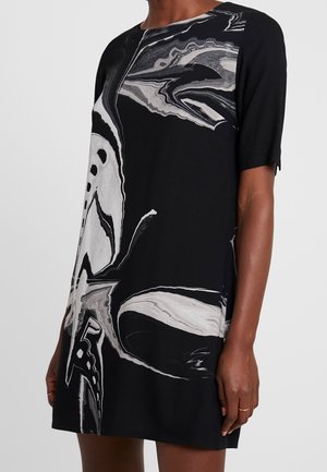 Robe noire à manches courtes avec motif abstrait de tourbillon marbré blanc et gris, portée par une personne à peau foncée avec des bagues dorées minimalistes aux doigts.