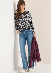 Langærmet sort-hvid mønstret top, lyseblå jeans, holder en burgunderfarvet jakke, hvide sneakers, står op ad en neutral væg.