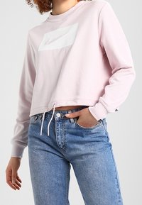 Růžový zkrácený svetr se bílým logem Nike, s dlouhými rukávy, šňůrkovým lemem a vhodný k svetrovým světle modrým džínsům s vysokým pasem.