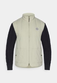 Μεγάλου παλτού με ανοιχτό μπεζ σώμα και σκούρες ναυτικές πλεχτές μανσέτες, κλείσιμο με φερμουάρ, ψηλό γιακά και στρογγυλό λογότυπο στο στήθος.