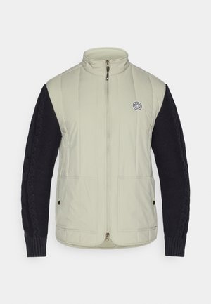 Jachetă căptușită cu un corp bej deschis și mâneci tricotate de culoare bleumarin închis, închidere cu fermoar, guler înalt și un logo rotund pe piept.