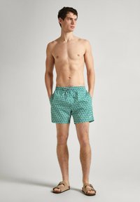 Turkio spalvos maudymosi šortai su balto lapų raštu, elastinga juosta ir raišteliu; modelis stovi basi, rankos kišenėse, torso nuogas.