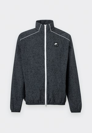 Μαύρο μπουφάν Nike Air με διακριτικό αφηρημένο σχέδιο, λευκό φερμουάρ, ενίσχυση στους ώμους και ελαστικά τελειώματα στα μανίκια και το κάτω μέρος.