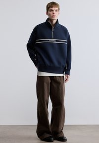 Φούτερ ναυτικού μπλε με φερμουάρ, λευκή ρίγα και λογότυπο στο στήθος, συνδυασμένο με καφέ παντελόνια φαρδιάς γραμμής και μαύρα παπούτσια. Λείο ύφασμα.