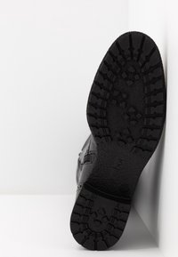 Scarpa in pelle nera con suola in gomma testurizzata, dotata di un battistrada decorato e di un logo. La scarpa è posizionata con la suola rivolta verso l'alto.