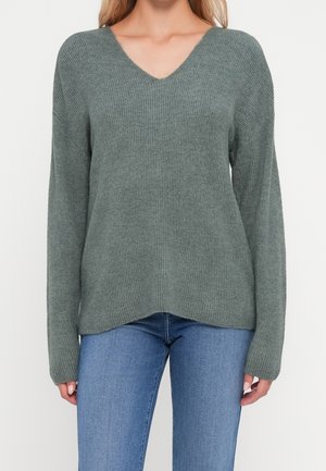 Femme portant un pull en tricot côtelé à manches longues de couleur vert pâle avec un col en V, assorti à un jean en denim bleu clair.