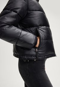 Giubbotto puffer nero con finitura lucida. Presenta sezioni trapuntate, una cerniera frontale e polsini a costine. Abbinato a jeans scuri.