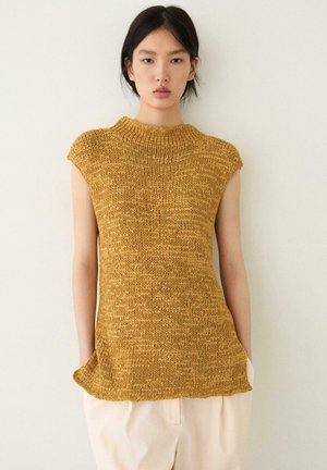 Frau trägt ein ärmelloses, senfgelbes, gestricktes Oberteil und weiße Hosen, steht mit den Händen in den Taschen vor einer schlichten hellen Wand.