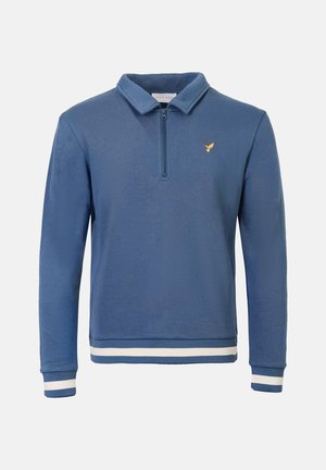 Mėlynas megztinis su puse užtrauktuko prie kaklo, kuriame yra mažas auksinis logotipas ant krūtinės ir baltos juostelės ant rankogalių bei apatinio krašto.