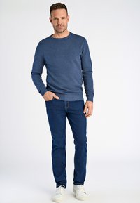 Maglione a maglia texturizzata blu con collo rotondo, abbinato a jeans scuri e sneaker bianche. Il modello si trova su uno sfondo neutro, con le mani in tasca.