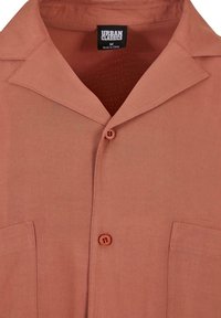 Camicia a maniche corte color ruggine con colletto da campo, taschino sul petto e due bottoni. Realizzata in tessuto liscio, presenta una sottile texture.