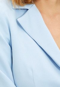 Blazer bleu clair en tissu texturé, avec des revers nets et des épaules structurées, présentant une coupe ajustée et des détails de couture subtils.