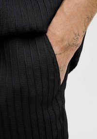 Sort tekstureret stof med lodrette striber, der har en lomme. Nærbillede viser hudfarve og en synlig tatovering på hånden.