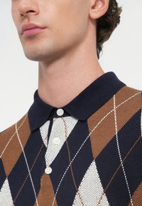 Strick-Polohemd mit einem marineblauen Kragen, einem weißen und braunen Rautenmuster und drei weißen Knöpfen am Ausschnitt.