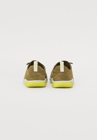 Pantofi din suede maro cu talpă albă și talpă texturată galbenă strălucitoare, având un tab de prindere în formă de buclă și un design minimalist.