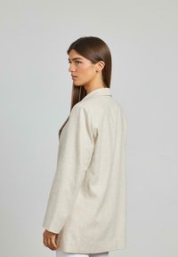 Blazer beige con tessuto testurizzato, vestibilità su misura e un bottone singolo. Presenta un colletto a punta e maniche lunghe, visto di lato.