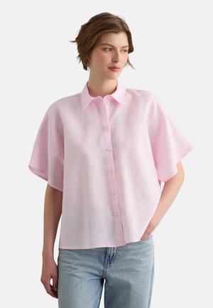 Vrouw met kort bruin haar, draagt een losse lichtroze korte mouw blouse met knopen en lichtblauwe jeans, hand in de zak, kijkt naar rechts.