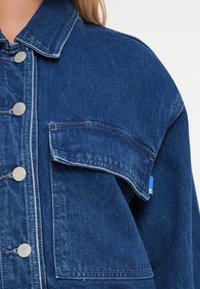 Jeans jakna u tamnoplavoj boji s ovratnikom, srebrnim kopčama i džepom na prsima. Ima kontrastni šav i oznaku s petljom.
