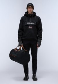 Sudadera de forro polar negra con un parche de logo y una bandera, combinada con pantalones negros y botas. El bolso de deporte presenta un logo y correas marrones.