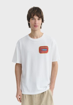 Mann trägt weißes T-Shirt mit rotem Sardinendosen-Motiv und beiger Hose, steht vor einem schlichten hellen Hintergrund.