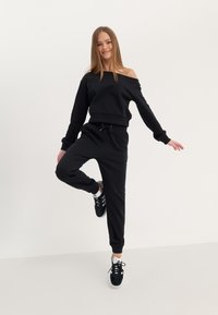 Sweat-shirt noir à design décolletté, avec des poignets élastiques, et un jogging noir assorti avec cordon de serrage. Des baskets noires complètent la tenue.