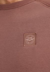 Sudadera de color óxido con un cuello de textura acanalada, que presenta un parche de tela cuadrado con un logo y un diseño alado.