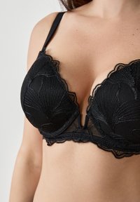Sujetador de encaje negro con borde ondulado, copas de malla texturizada con patrones florales y tirantes ajustables. Detalle central en forma de V.