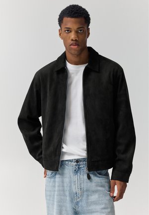 Bărbat tânăr purtând o jachetă neagră cu fermoar peste un tricou alb și blugi deschiși la culoare, stând în picioare în fața unui fundal simplu și deschis la culoare.