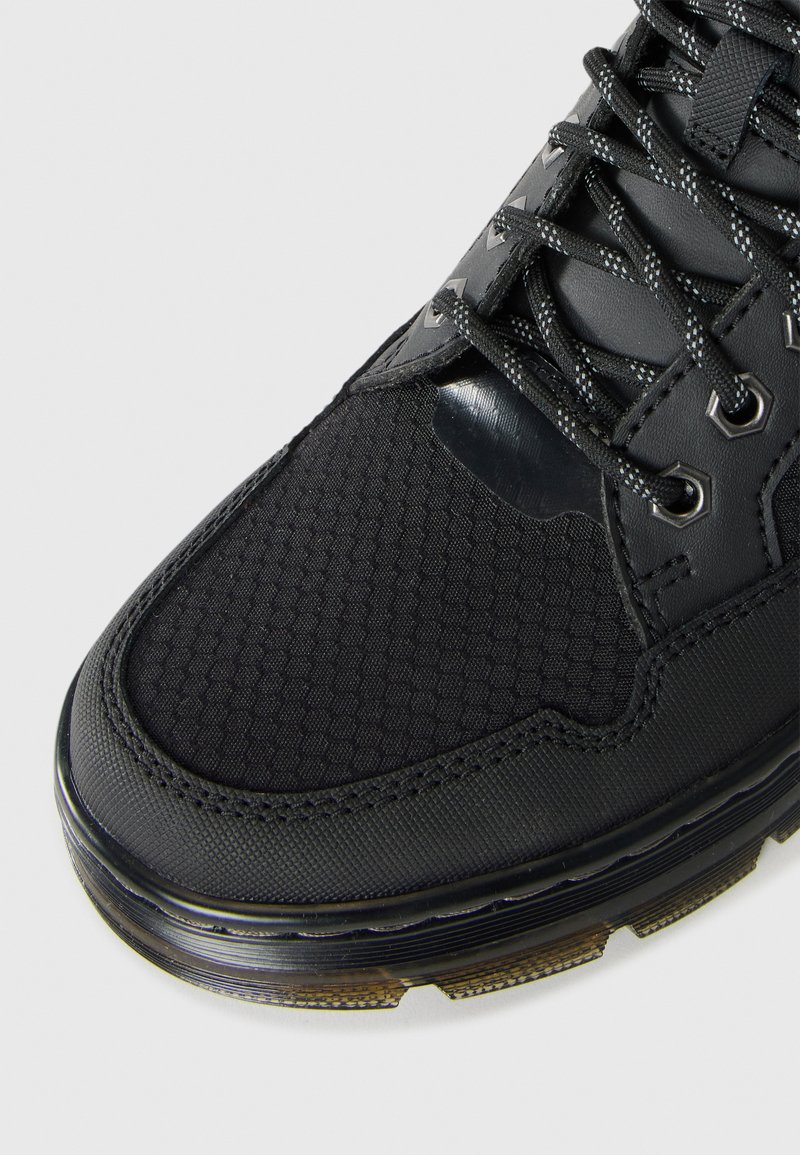 Détail d'une chaussure de sport noire montrant un bout en tissu texturé, des coutures renforcées, des lacets noirs à motif et une semelle en caoutchouc translucide.