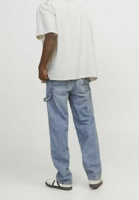 Lichtkleurig kort mouw shirt, blauwe losse jeans met multi-zakken ontwerp en sandalen met zwart-witte banden. Achteraanzicht.