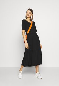 Svart bomull midi-kjole med kortermede, lagvis volangkant og rund hals. Kombinert med hvite joggesko og en oransje crossover-veske.